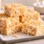 Stacked homemade Rice Krispie treats on a tray with marshmallows and butter in a bright kitchen.